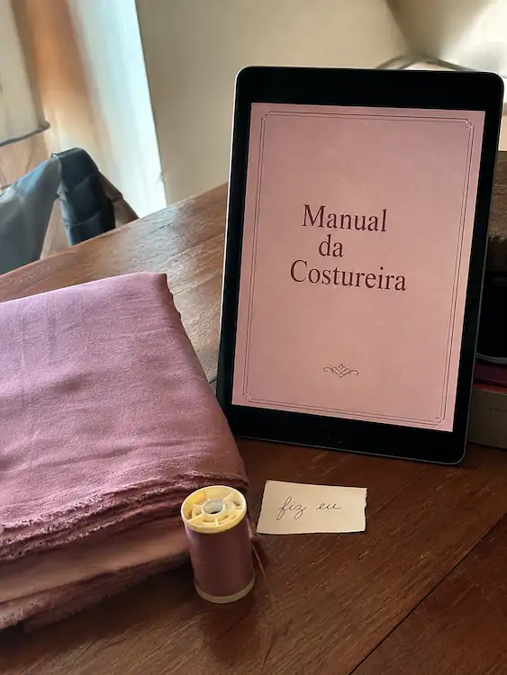 Tablet mostrando a capa do Manual da Costureira, apoiado sobre uma mesa de madeira com tecido rosa dobrado, carretel de linha e cartão escrito 'fiz eu'